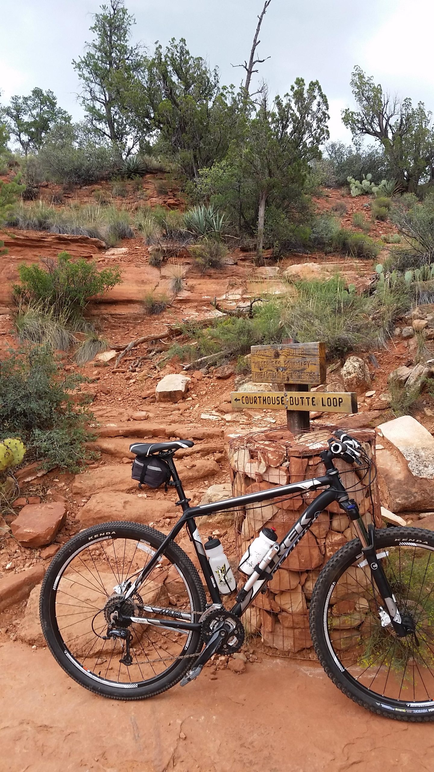 Mountain bike positioned beside a wooden trail sign indicating the "Courthouse Butte Loop," surrounded by reddish rock formations and desert vegetation. Bell Rock Trailway mountain bike trail.