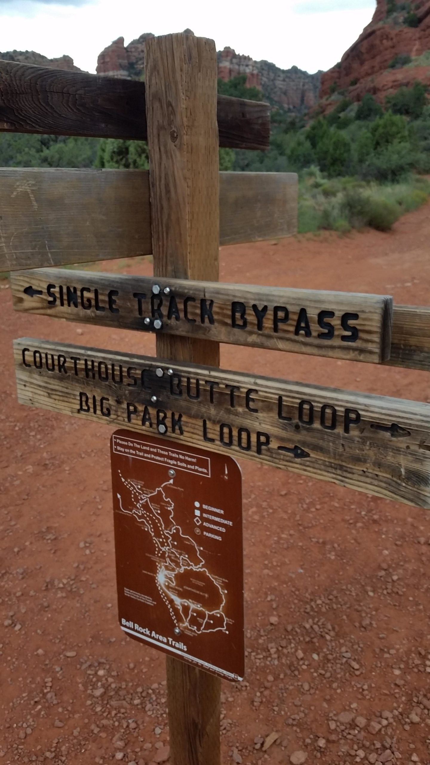 Wooden trail sign with directions for hiking paths, including "Single Track Bypass," "Courthouse Butte Loop," and "Big Park Loop," along with a map of the Bell Rock Area Trails indicating difficulty levels. The background features red rock formations and greenery, typical of a scenic outdoor landscape. Bell Rock Trailway mountain bike trail.