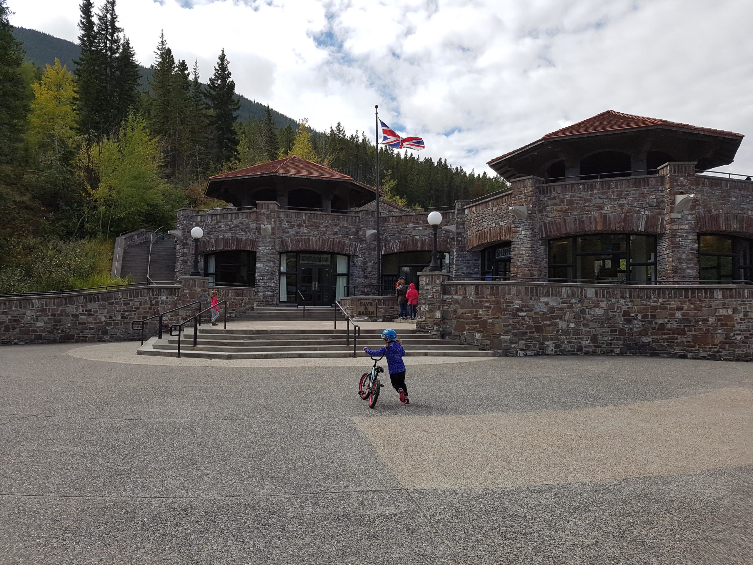 A child wearing a blue helmet and jacket rides a small bicycle in front of a stone building with a tiled roof. The building features large windows and is surrounded by trees. A flag is flying above the entrance, and other visitors can be seen approaching the building. The sky is partly cloudy, and the area appears to be a recreational or visitor center. Sundance mountain bike trail.