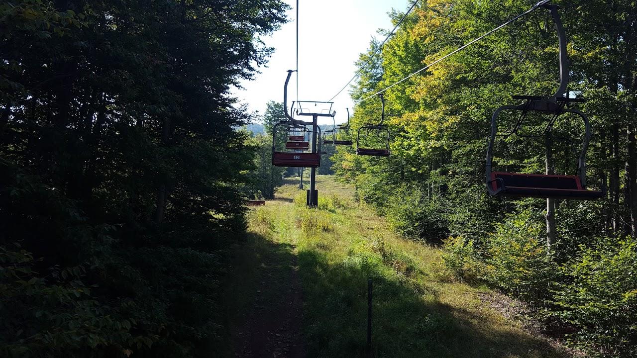 A scenic view of a ski lift with empty red chairs, surrounded by lush green trees and a clear blue sky in the background. The path below is lined with grass and shrubs, indicating a tranquil outdoor setting. Timberline mountain bike trail.