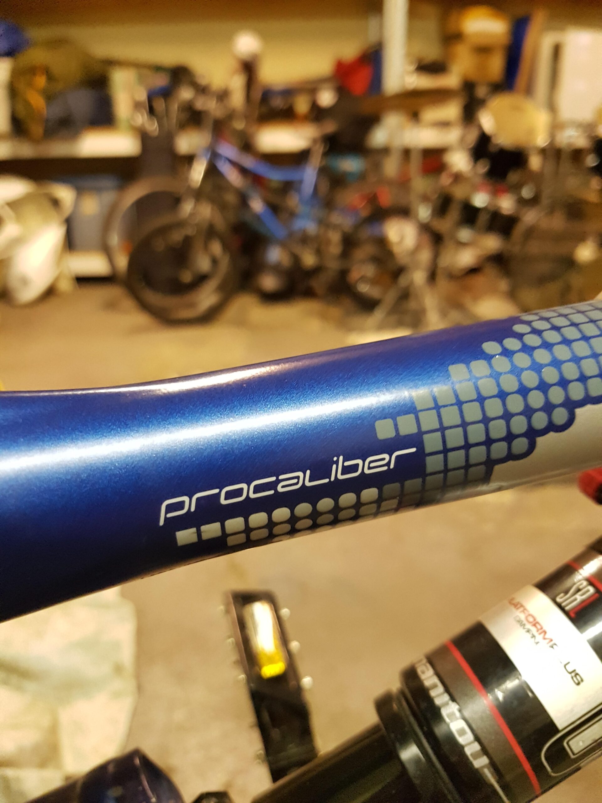 Gary Fisher PRO: Close-up of a bike frame with the word "Procaliber" prominently displayed, set against a blurred background of various bicycles and garage items. The frame features a sleek blue finish with a modern graphic design.