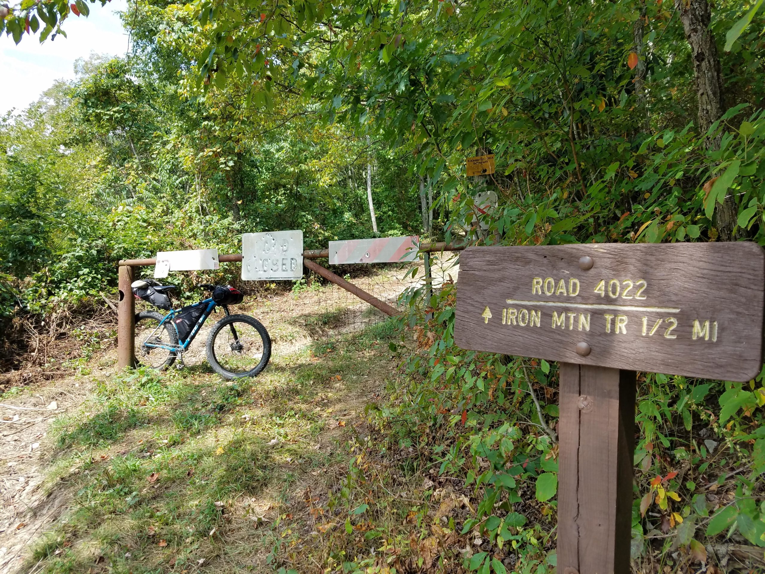 A bicycle leaning against a gate with a "Road Closed" sign, surrounded by greenery. A wooden sign indicates "Road 4022" and points to the "Iron Mountain Trail 1/2 Mile." The scene captures a tranquil, wooded area with trees and shrubs. Iron Mountain mountain bike trail.