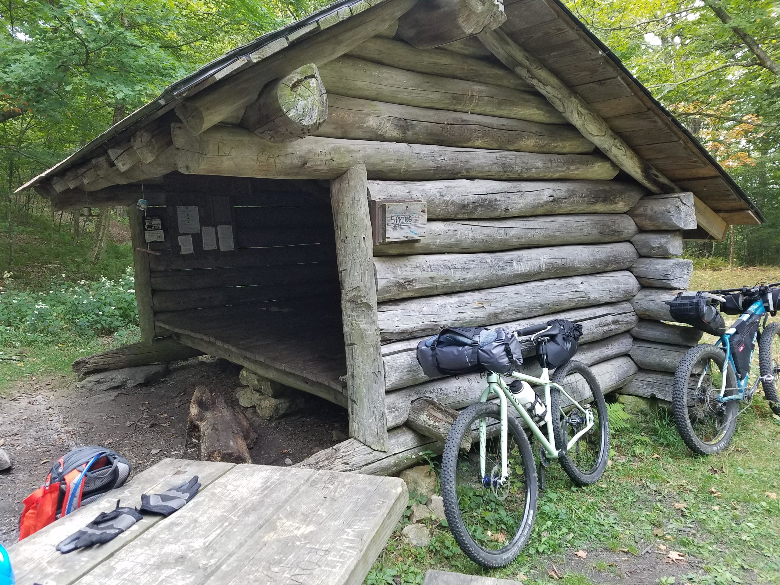 A rustic log cabin shelter in a forested area, with two bicycles equipped with saddlebags parked nearby. A wooden picnic table is visible in the foreground, along with a pair of gloves and a backpack. The surroundings feature green foliage and wildflowers, contributing to a serene outdoor setting. Iron Mountain mountain bike trail.