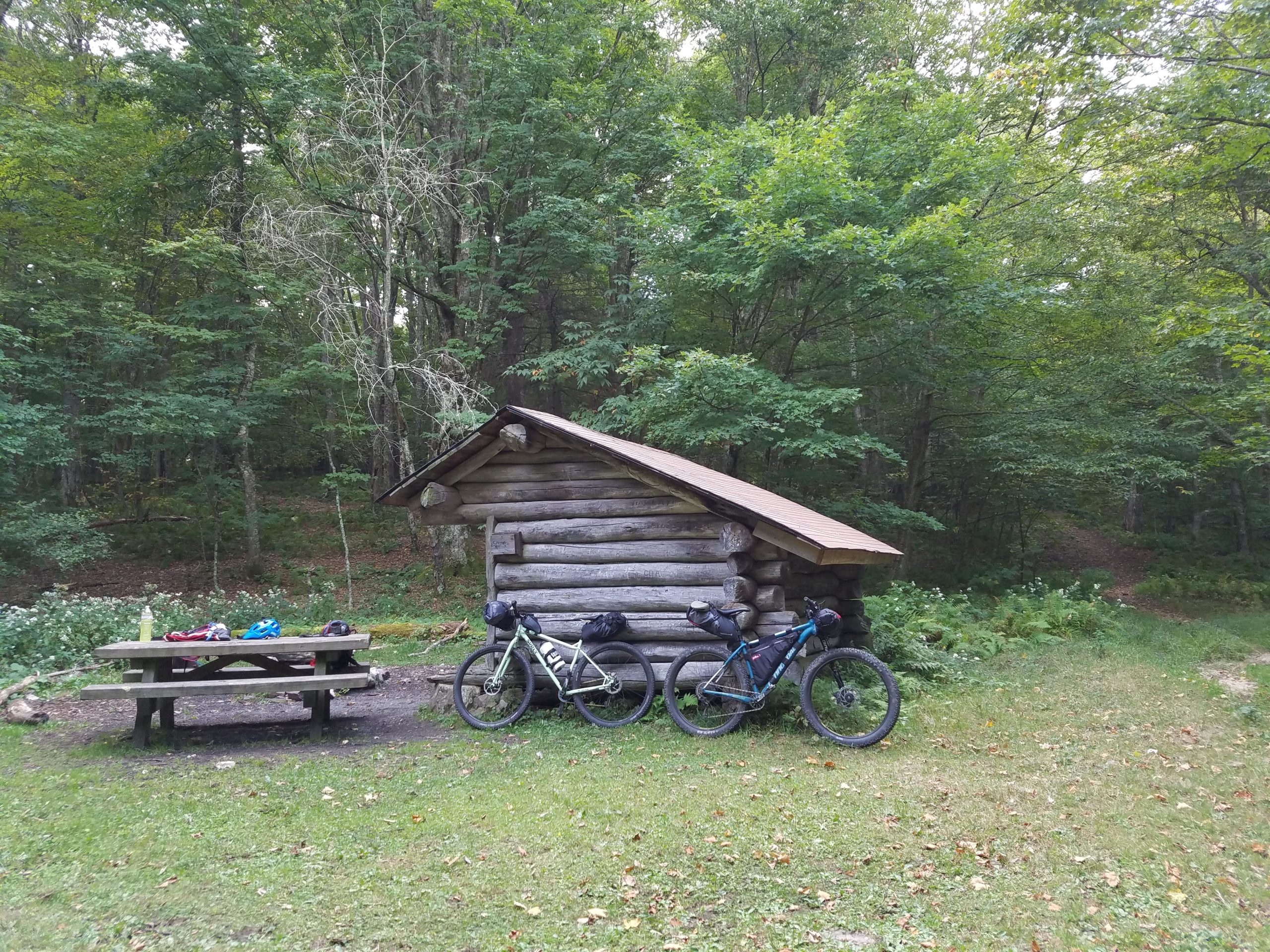 A rustic log cabin surrounded by trees, with two bicycles parked nearby and a picnic table in the foreground. Helmets and gear are placed on the table, suggesting an outdoor cycling stop in a natural setting. Iron Mountain mountain bike trail.