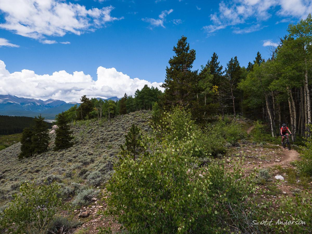 A mountain biker rides along a winding, rocky trail surrounded by lush greenery and trees, with rolling hills in the background under a partly cloudy blue sky. Colorado Mountain College Trails mountain bike trail.