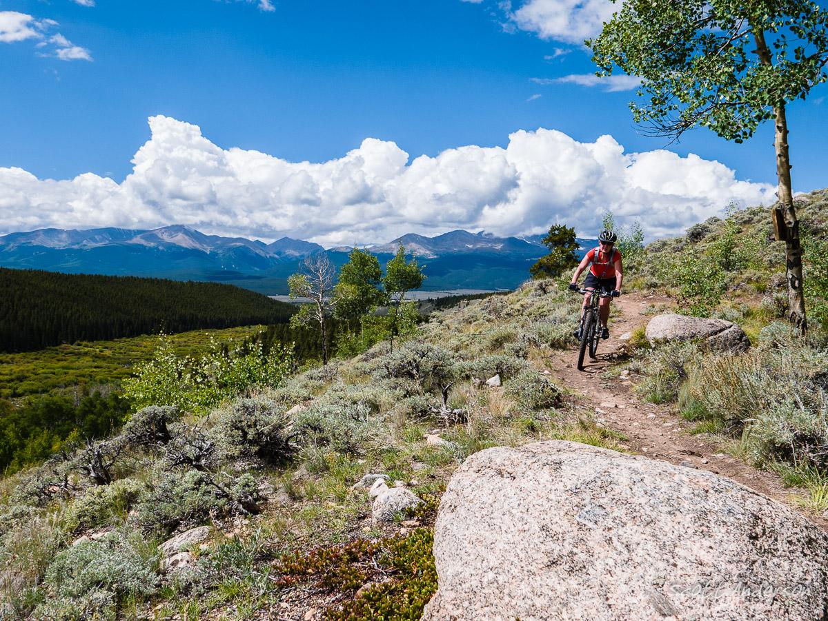 A mountain biker rides along a rocky trail surrounded by lush greenery and rolling hills, under a bright blue sky dotted with white clouds and mountain peaks in the background. Colorado Mountain College Trails mountain bike trail.