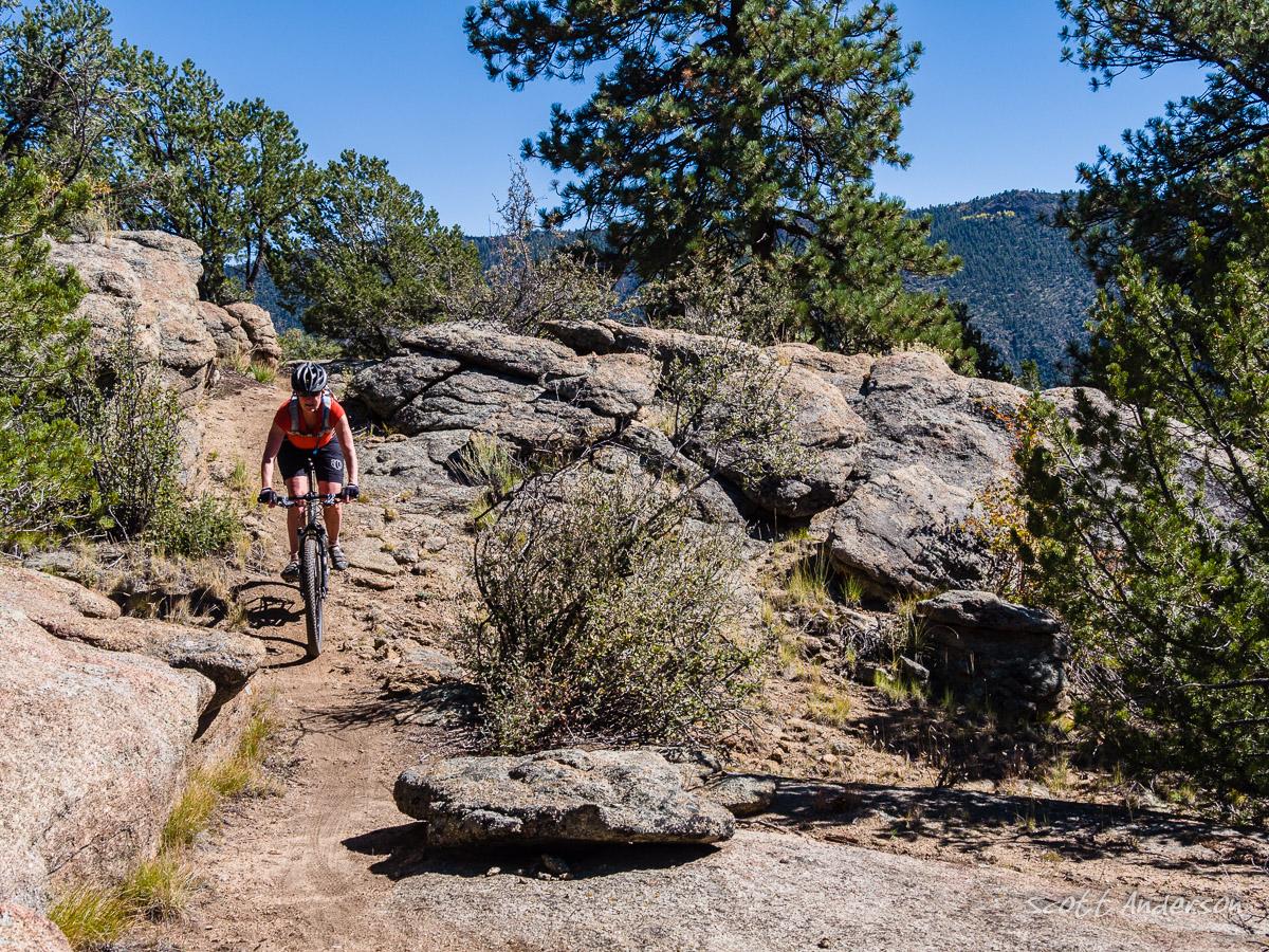A mountain biker navigates a rocky trail surrounded by pine trees and mountains under a clear blue sky. The terrain features large boulders and patches of grass, showcasing a rugged outdoor environment. Midland Hills Trails mountain bike trail.