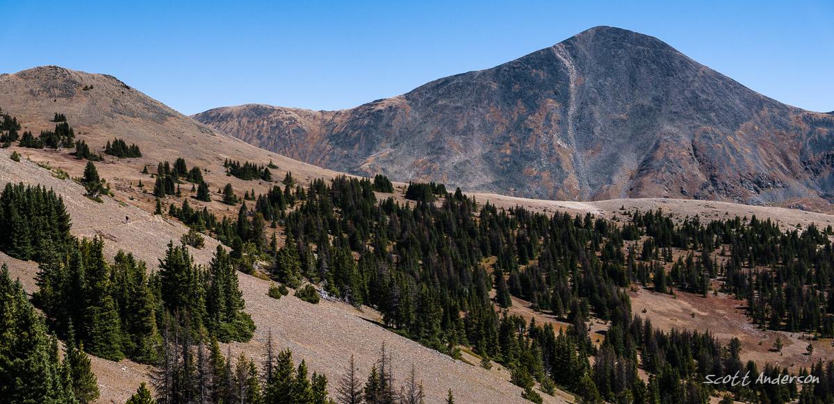 A scenic view of mountains with a prominent peak in the background. The foreground features rolling hills covered with sparse vegetation and patches of evergreen trees, under a clear blue sky. The landscape showcases a mix of rocky terrain and lush greenery, highlighting the natural beauty of the mountainous region. Monarch Crest Trail mountain bike trail.