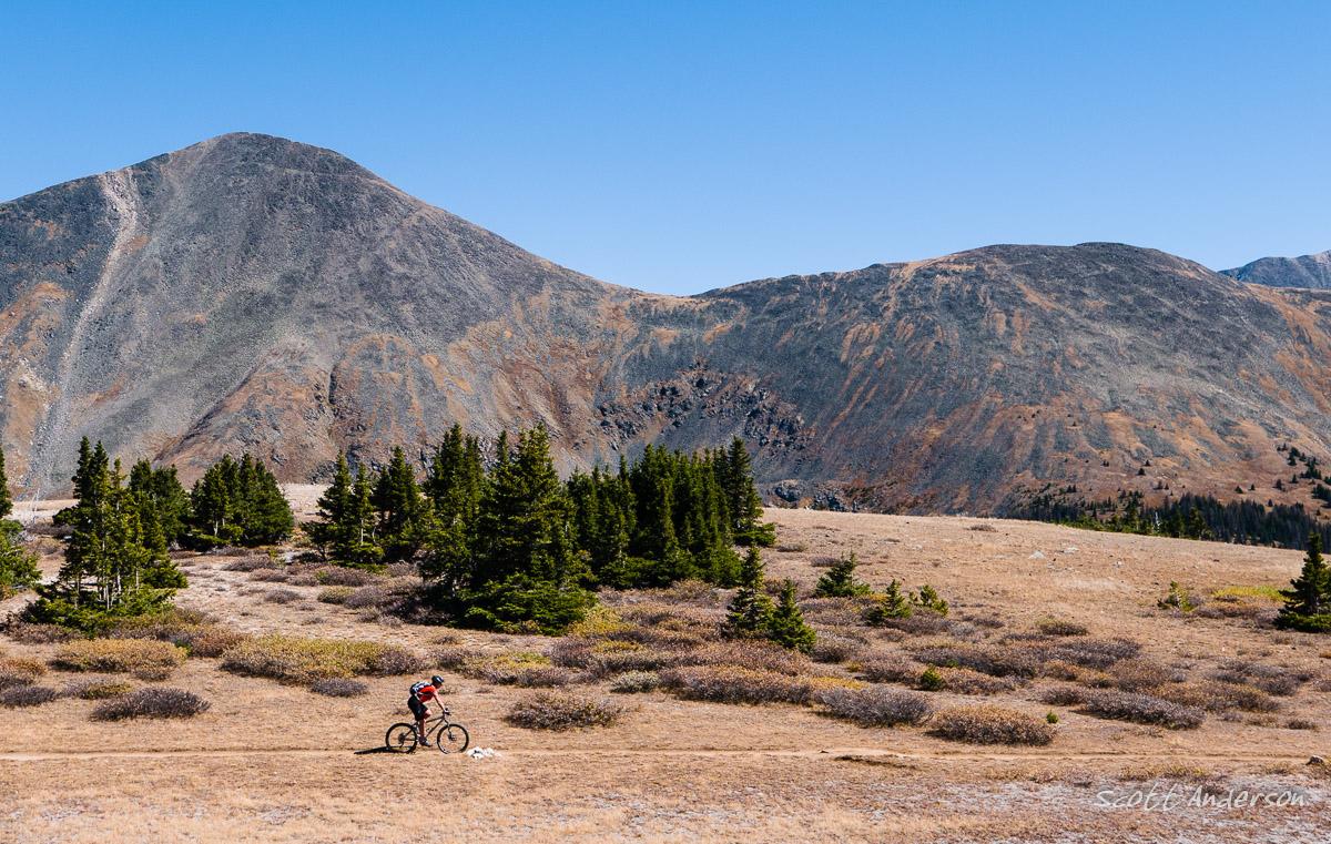 A mountain biker rides along a dirt path through an expansive, rugged landscape featuring rolling hills, patches of greenery, and sparse vegetation under a clear blue sky. The backdrop includes prominent mountain peaks, showcasing earthy tones and textures. Monarch Crest Trail mountain bike trail.