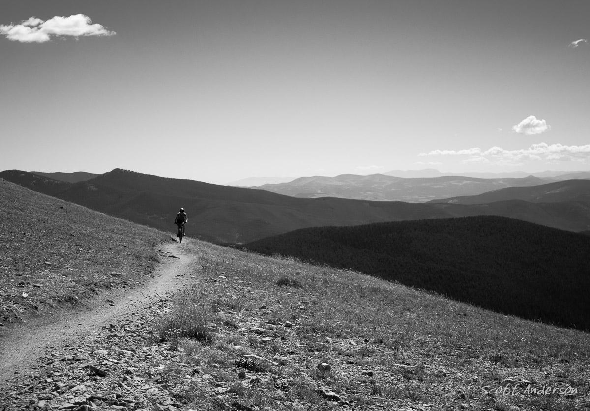 A lone mountain biker rides along a winding dirt path on a hillside, surrounded by expansive mountain views under a clear sky. The image is in black and white, highlighting the natural textures of the terrain and the silhouette of the rider against the distant mountains. Monarch Crest Trail mountain bike trail.