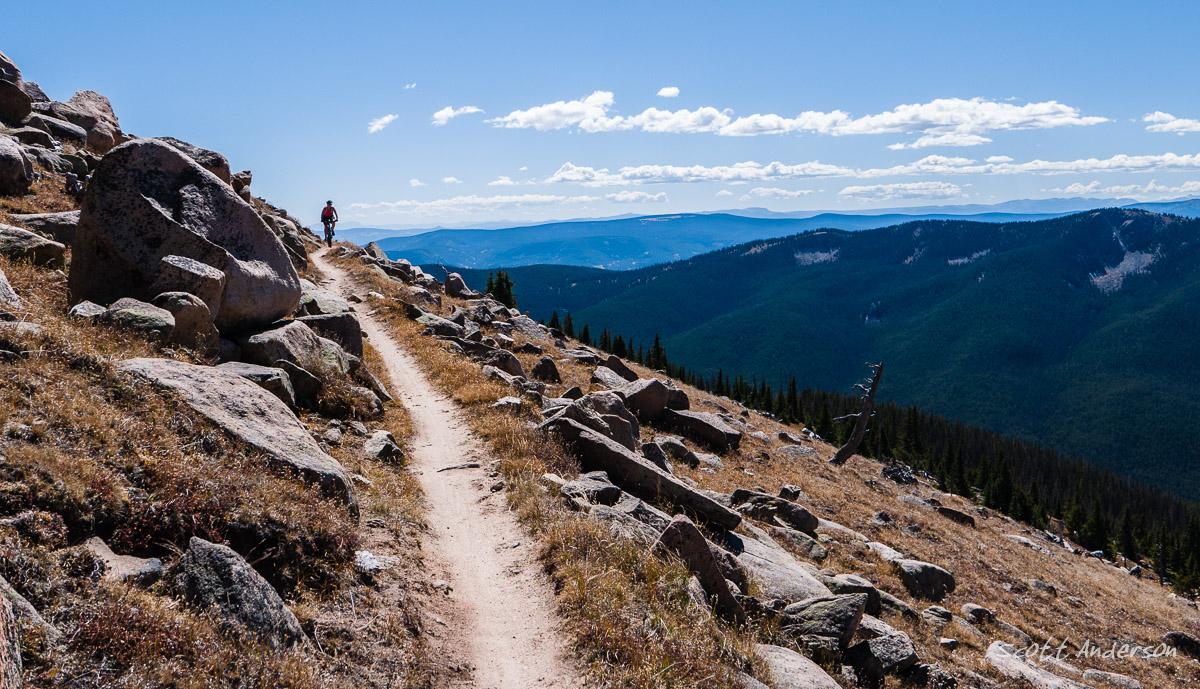 A hiker walking along a narrow dirt path that winds through rocky terrain, with expansive views of rolling mountains and a clear blue sky in the background. The landscape is dotted with patches of grass and trees. Monarch Crest Trail mountain bike trail.