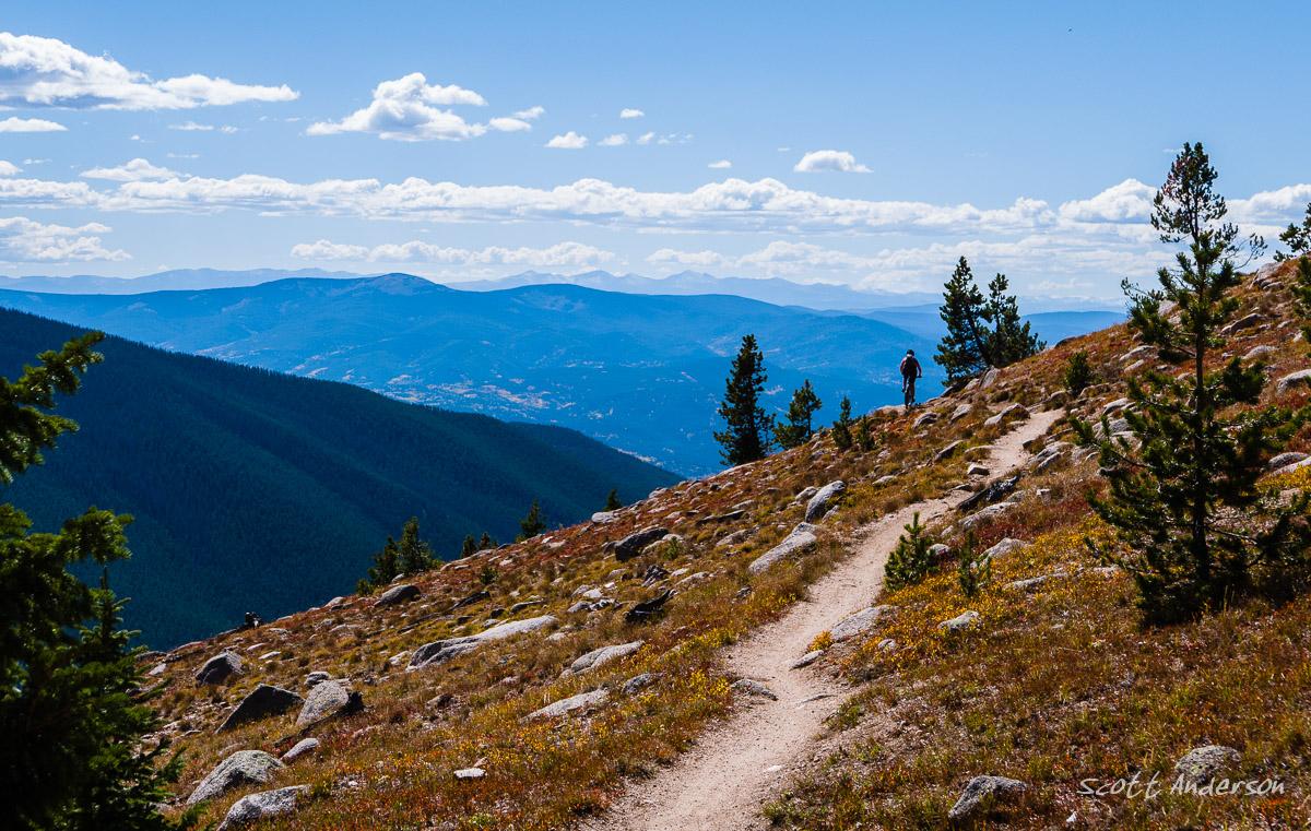 A hiker walking along a winding trail on a mountain slope, surrounded by rocky terrain and evergreen trees. In the background, expansive mountain ranges and a clear blue sky with scattered clouds. Monarch Crest Trail mountain bike trail.