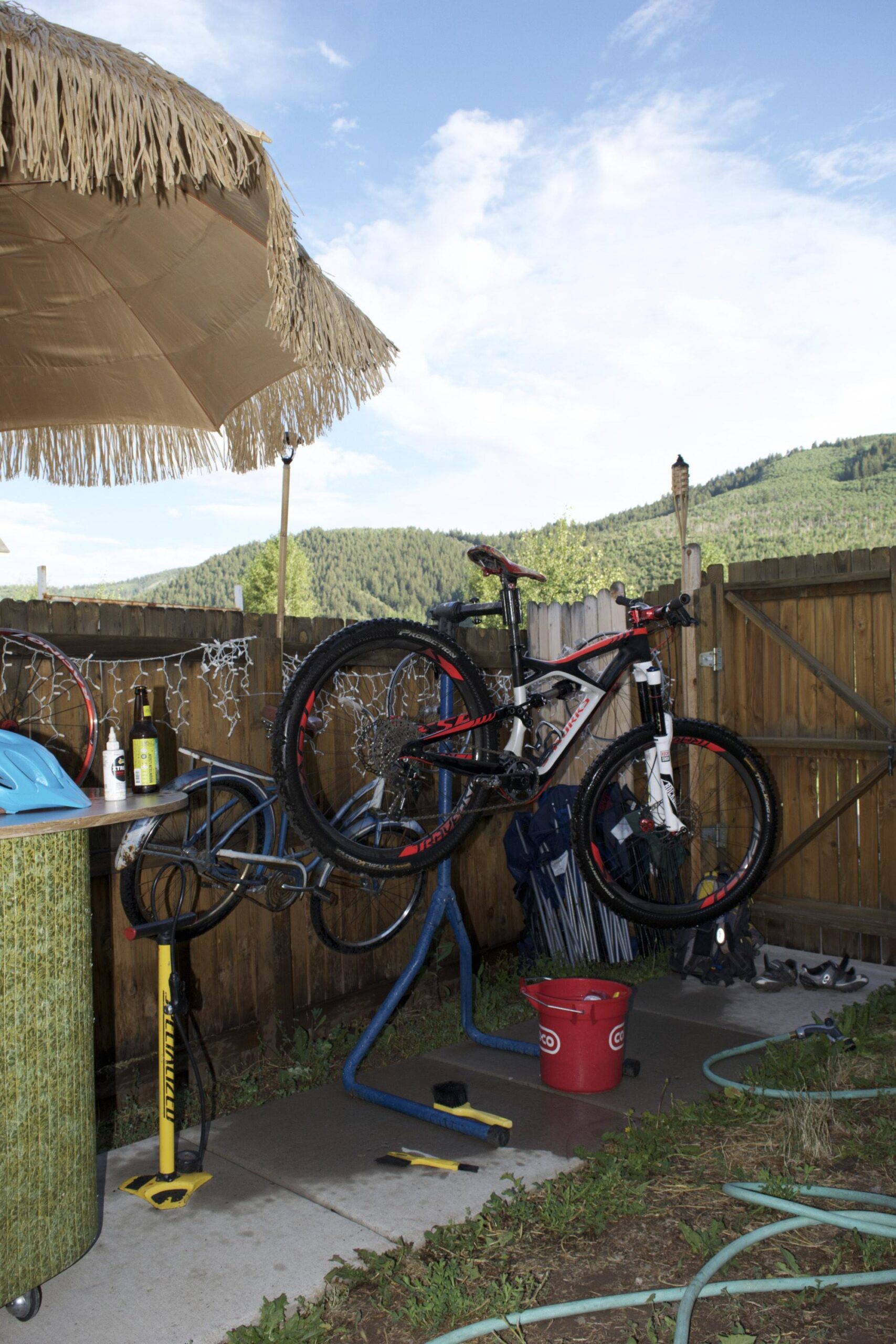 Specialized Enduro S-Works 650b: A backyard area with a shaded umbrella and two mountain bikes mounted on a repair stand. A small table with drinks, a bicycle pump, and a red bucket are visible, along with tools and gardening supplies scattered on the ground. The backdrop features a wooden fence and a hillside landscape under a partly cloudy sky.