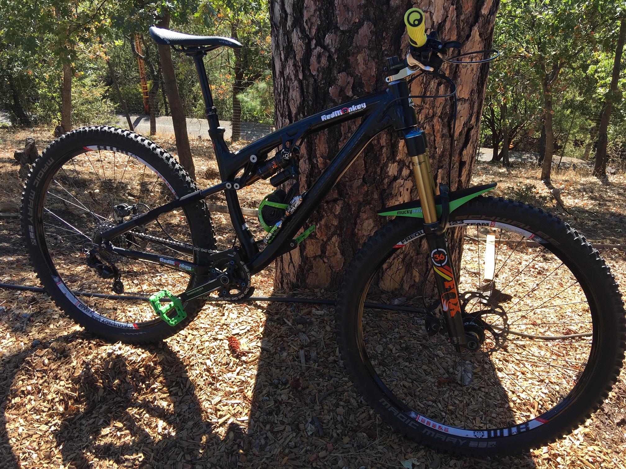 Rocky Mountain Instinct 970 MSL: A black mountain bike with green pedals and a gold front fork, positioned next to a tree in a natural setting with dry leaves and wood chips on the ground.