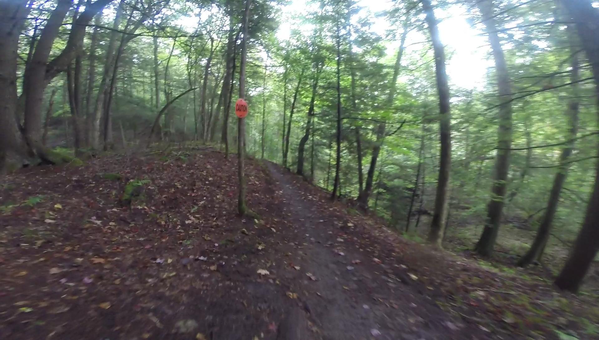 A winding dirt trail through a forest, surrounded by tall trees and lush greenery. A caution sign is visible beside the path, which is covered in fallen leaves and light mist. The atmosphere appears serene and inviting, suggesting a peaceful outdoor setting. Kingdom Trails mountain bike trail.