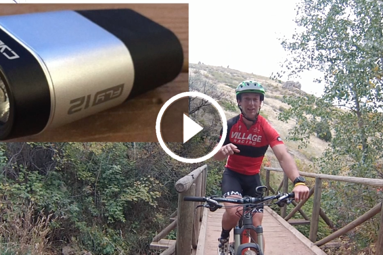 A split image featuring a close-up of a cycling camera on the left and a cyclist wearing a red jersey and helmet on the right, riding a mountain bike across a wooden bridge surrounded by greenery. A play button icon indicates a video element.