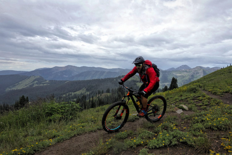 A mountain biker in a red jacket and helmet rides along a narrow dirt path through lush green hills, surrounded by scenic mountain views under a cloudy sky. Wildflowers dot the landscape as the biker navigates the terrain.