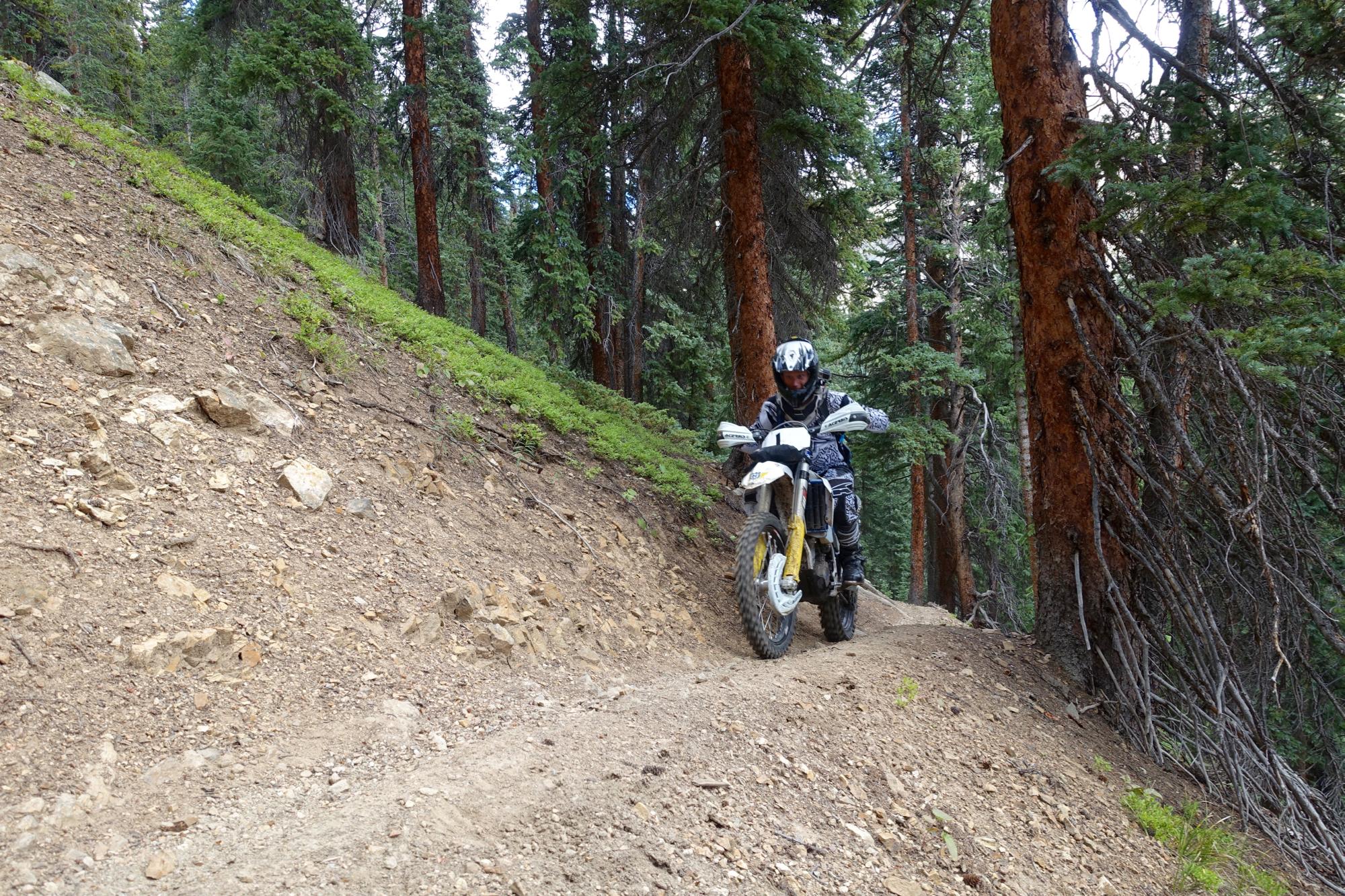 A rider in protective gear maneuvers a dirt bike along a narrow, rocky trail surrounded by tall trees and greenery. Timberline Trail mountain bike trail.