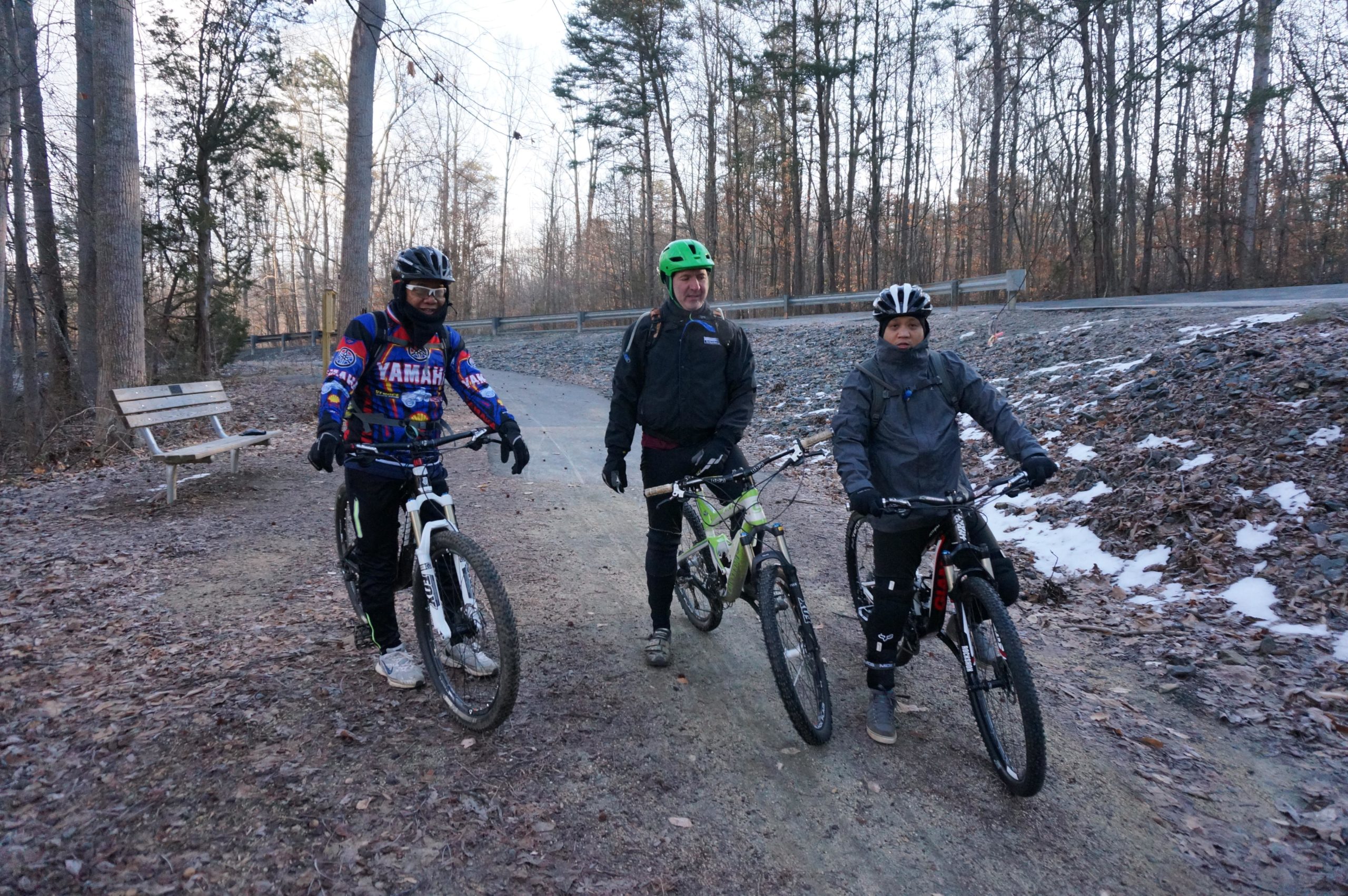 Three people stand with mountain bikes along a dirt path in a wooded area. The individuals are wearing cycling gear, including helmets. A wooden bench is visible in the background, surrounded by bare trees and scattered leaves, indicating a cool season. The scene suggests a recreational outing or biking adventure. Salem Lake mountain bike trail.