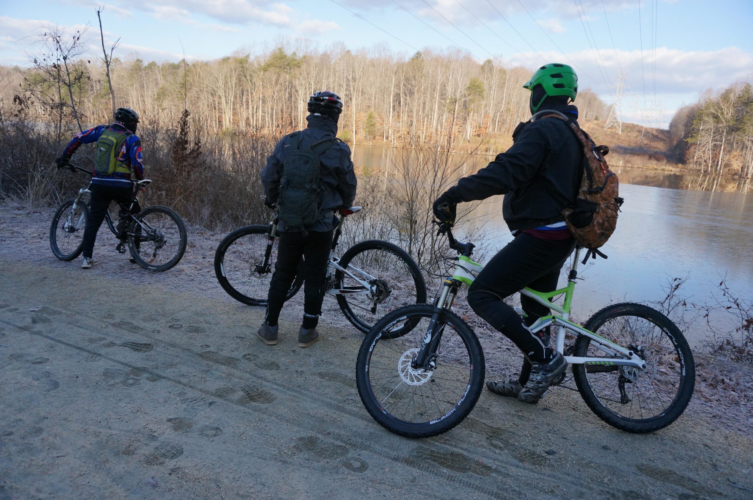Three mountain bikers pause on a dirt path near a frozen lake, surrounded by bare trees and a serene winter landscape. One biker, wearing a colorful jacket and backpack, stands facing the water, while the others look out towards the lake. The scene captures a moment of outdoor adventure in a tranquil setting. Salem Lake mountain bike trail.