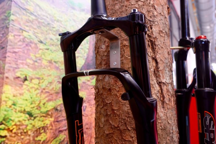 A close-up view of a black bicycle fork mounted beside a tree trunk, with a blurred natural landscape background. The fork features sleek design elements and is attached to a metal bracket.