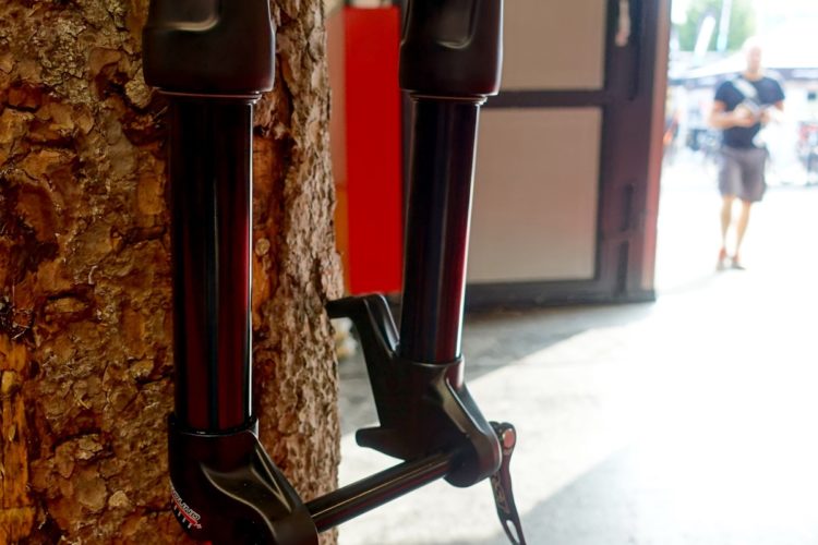 A close-up view of a bicycle fork mounted on a tree trunk, with a blurred background showing a person walking out of a workshop or garage. The fork has a shiny black finish and is designed for suspension.
