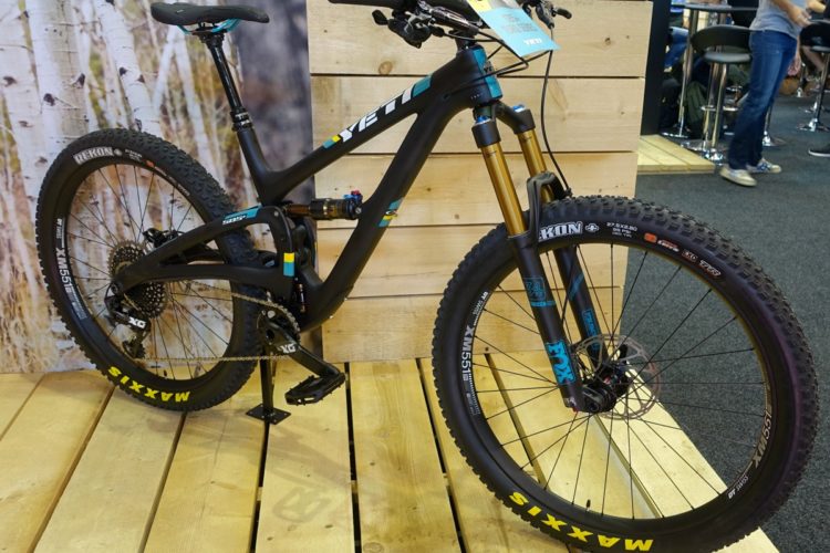 Alt text: A black mountain bike from Yeti, featuring a sleek design with turquoise and yellow accents, standing on a wooden platform. The bike has prominent Maxxis tires and is equipped with a front suspension fork. In the background, there are blurred figures and a wooden panel, suggesting a trade show or exhibition setting.