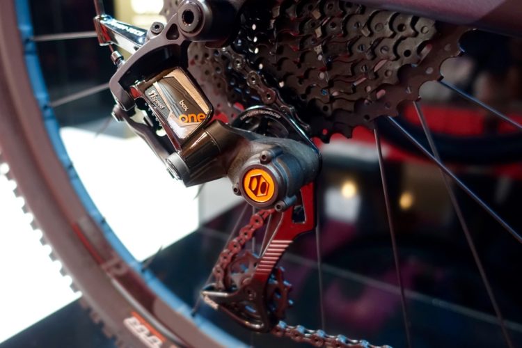 An image of a bicycle's rear derailleur and cassette, showcasing intricate details of the components. The derailleur features a shiny metallic casing with the label "box one," and an orange-highlighted gear mechanism. The black chain and gears are clearly visible, set against a blurred background.