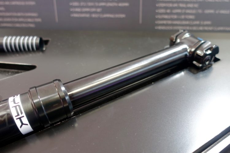 Close-up view of a black bicycle seat post, featuring a cylindrical design with a visible clamp mechanism at one end. The background is dark with visible textures and lines, suggesting a workspace or display area for bike components.