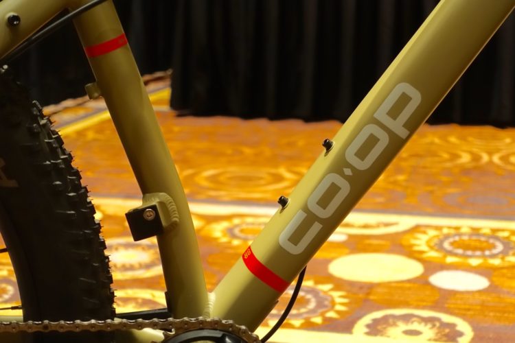 Close-up view of a mountain bike frame, featuring a muted olive green color with a prominent "co-op" logo in white. The bike has a thick, knobby tire and a red accent on the frame. The background shows a patterned carpet, suggesting an event or exhibition setting.
