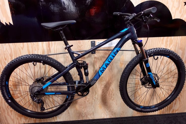 A Marin mountain bike displayed against a wooden background. The bike features a sleek black frame with blue accents, large knobby tires, and a dual suspension system, showcasing its off-road capabilities.