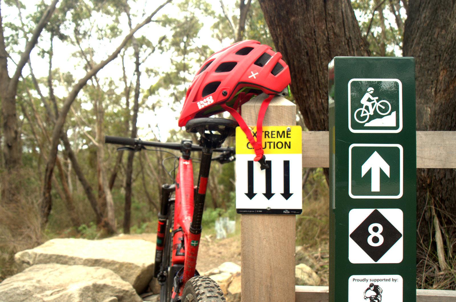 A red bicycle helmet rests on the handlebars of a mountain bike, positioned next to trail signage. The signs indicate "Extreme Caution" with arrows pointing downward and provide directions for cyclists, including symbols for biking paths and difficulty ratings. The background features a wooded area with trees, creating a natural, outdoor setting. Arthurs Seat MTB Park mountain bike trail.