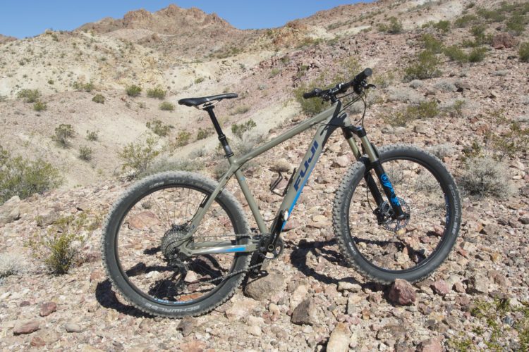 A mountain bike on rocky terrain in a desert landscape, featuring a grey frame with blue accents and wide tires. The background includes rugged hills and sparse vegetation, under a clear blue sky.