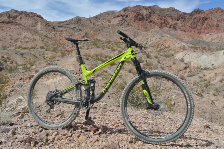 A vibrant green and black mountain bike is positioned on rocky terrain with desert hills in the background under a partly cloudy sky. The bike features knobby tires suitable for off-road trails and a sleek frame design.