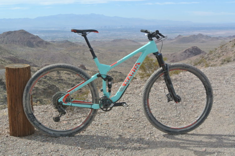 A turquoise mountain bike resting against a wooden post, set against a background of rugged desert terrain and distant mountains. The bike features black and red accents, thick tires, and a visible gear system, with a panoramic view of a valley in the background under a clear sky.