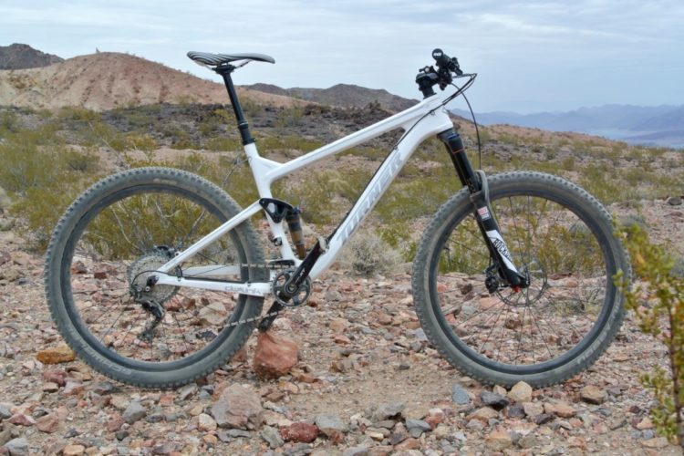 A white mountain bike parked on rocky terrain with sparse vegetation and distant hills in the background. The bike features thick tires, a dual-suspension system, and a sleek design, suggesting it is built for off-road adventures. The sky is cloudy, reflecting an overcast day.