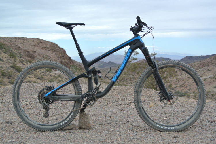 A mountain bike with a black and blue frame is parked on rocky terrain, surrounded by hills and a cloudy sky in the background. The bike features thick, knobby tires and a modern design, suggesting it's suited for rugged trails.
