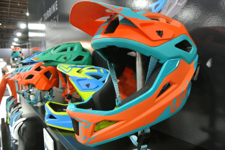 A close-up view of brightly colored bike helmets displayed on a shelf, featuring a variety of designs in orange, turquoise, green, and yellow. The helmets are designed for safety and style, with ventilation openings and adjustable straps.