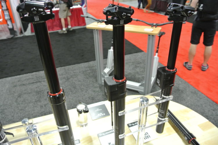 Three black bicycle dropper posts on display at an outdoor equipment trade show, featuring various components and markings. In the background, a blurred view of attendees and displays can be seen on a red and black carpet.