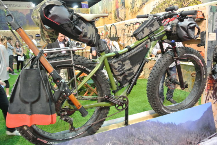 A customized fat bike with camouflage bags attached for storage, featuring a shovel and various accessories. The bike is displayed against a colorful backdrop, with people and outdoor gear visible in the background.