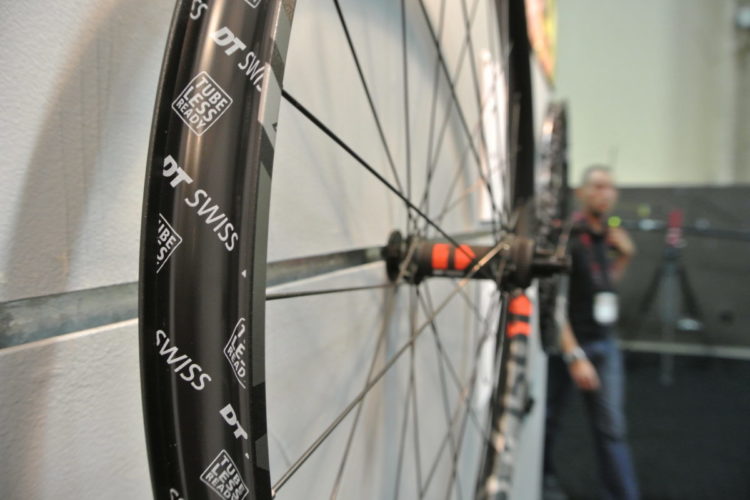 A close-up view of a black bicycle wheel featuring the "DT Swiss" logo, mounted on a display. The wheel showcases a tubeless-ready design with visible spokes and a hub, while a person is partially blurred in the background.