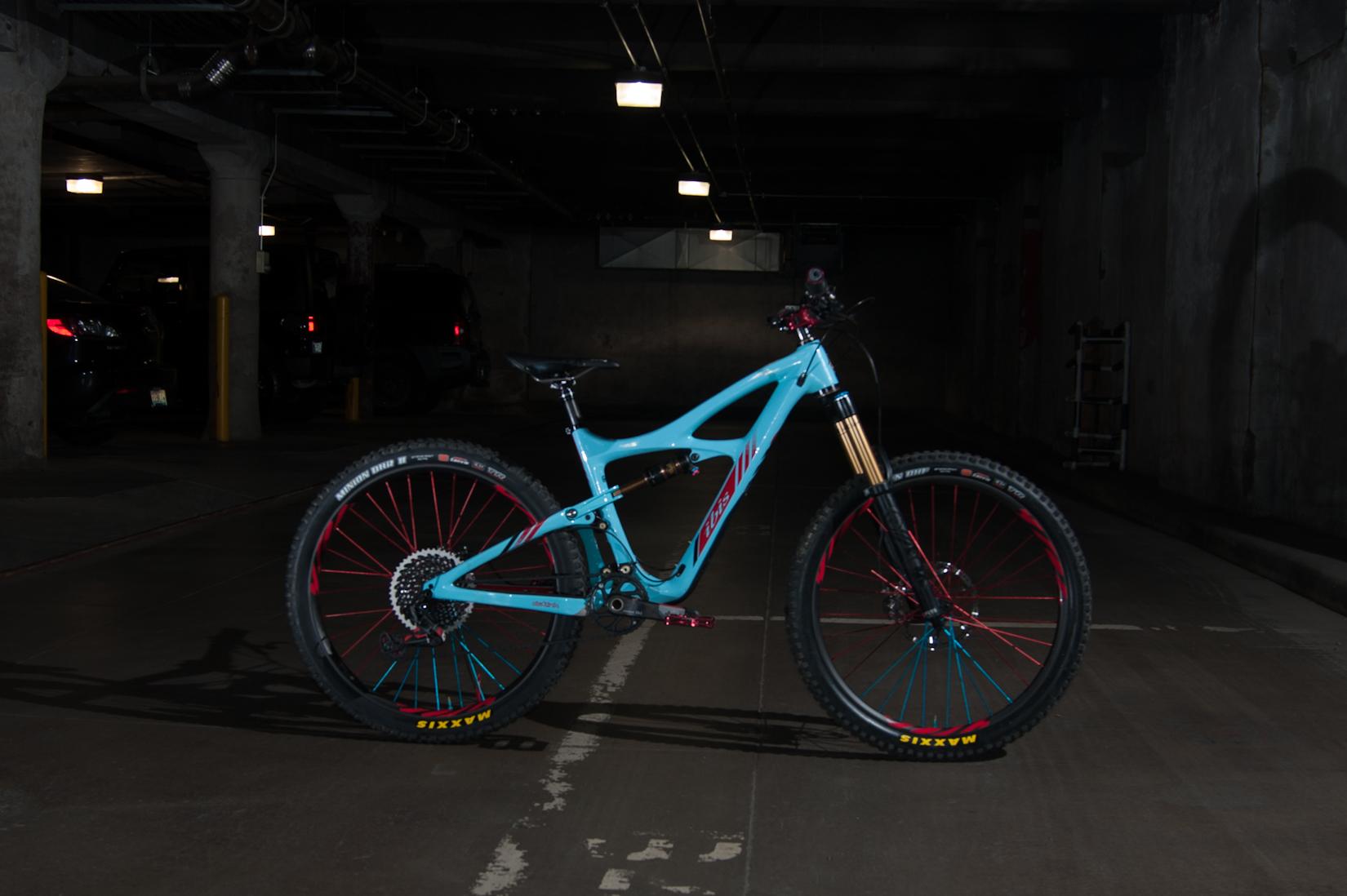 Ibis Mojo HD3: A blue mountain bike with red accents and black tires, positioned in a dimly lit parking garage. The bike features a full suspension frame and is centered on a concrete surface with tire marks, while cars are faintly visible in the background.