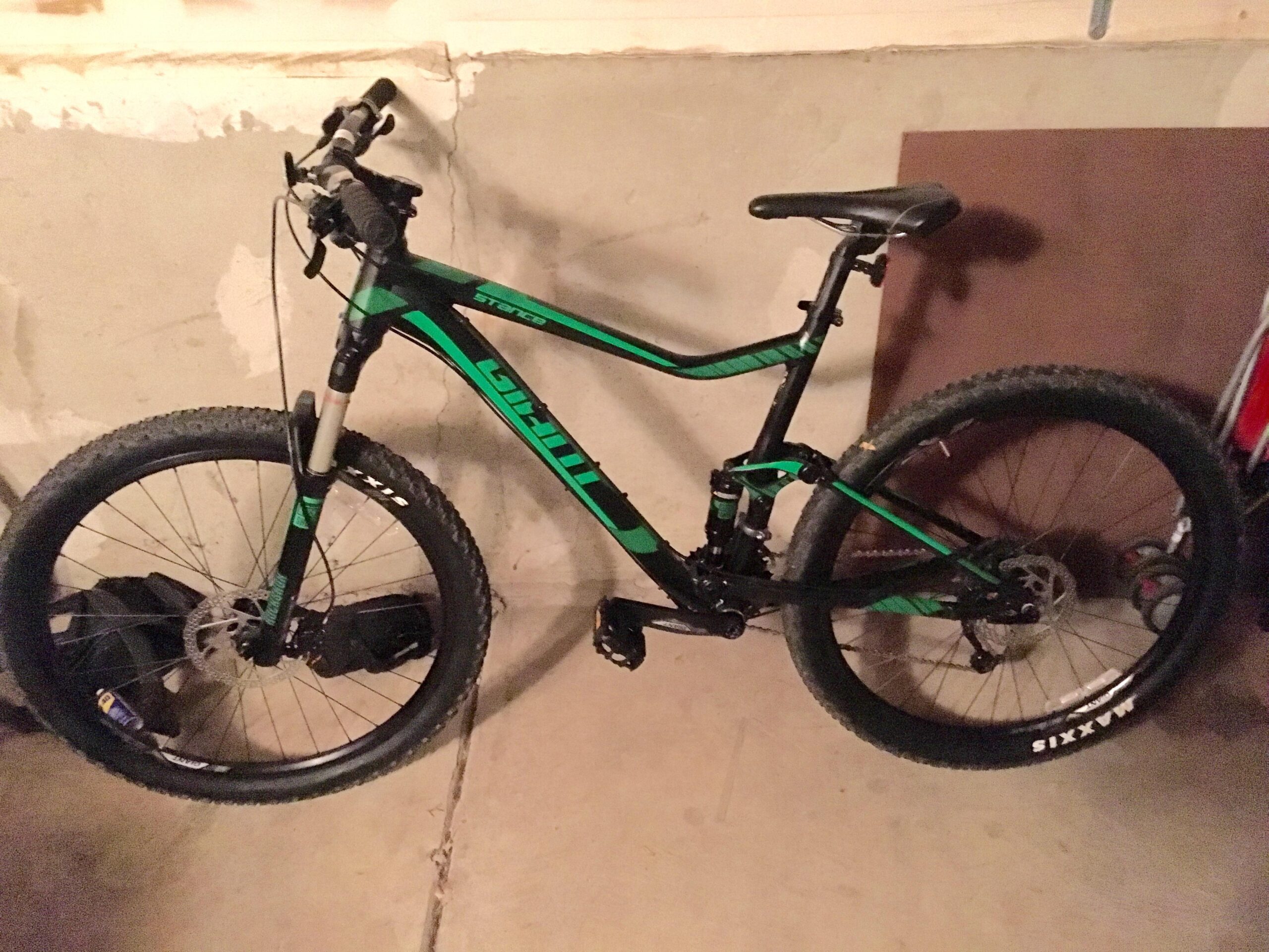 Giant Stance 27.5 2: A green and black mountain bike is parked in a garage, leaning against a wall. The bike features thick tires, a suspension fork, and a dual suspension frame. The garage floor is concrete, and there is some equipment visible in the background.