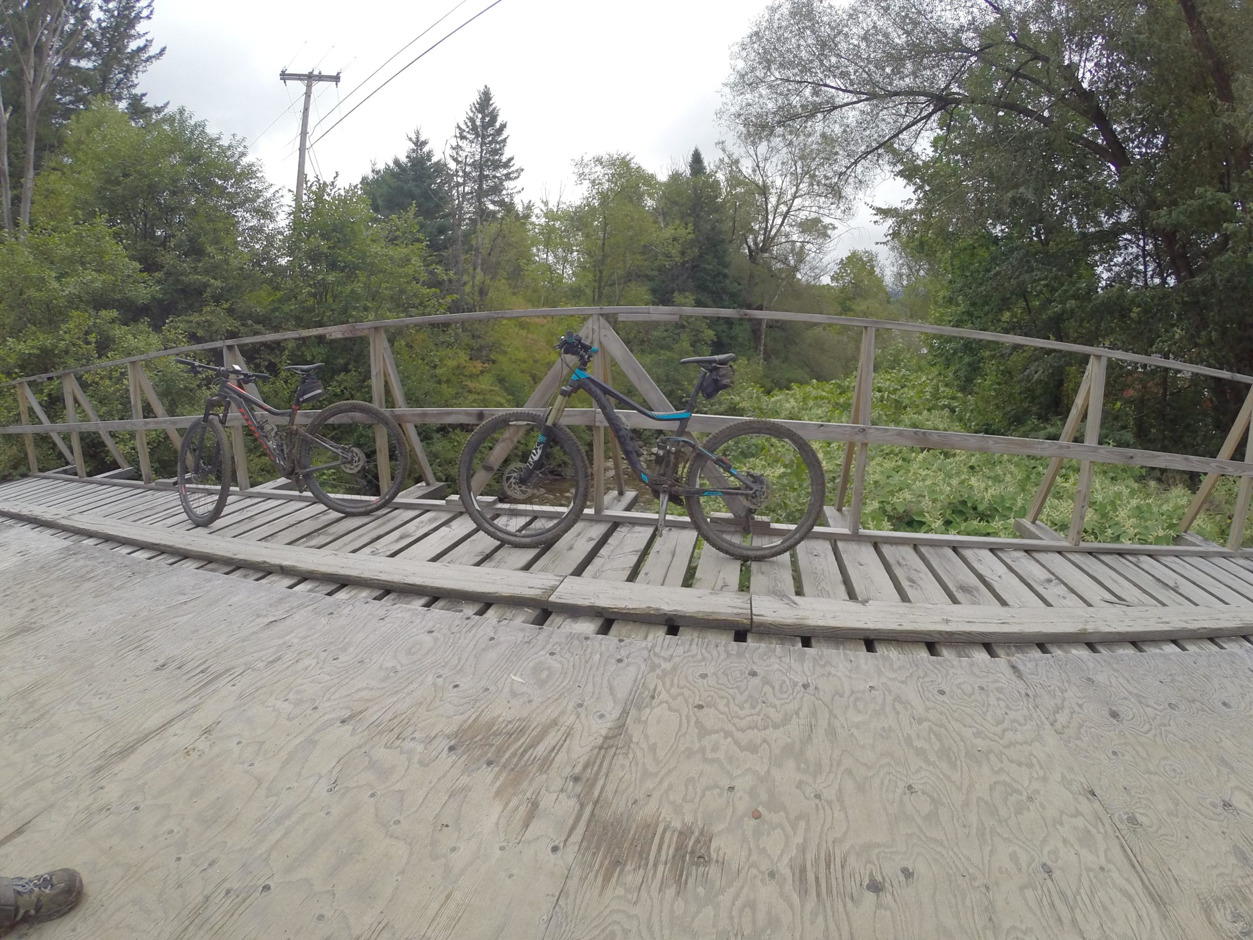 Two mountain bikes with dirt on them are parked on a wooden bridge surrounded by greenery. The bridge has a slight curve and is set in a natural outdoor environment with trees in the background. Kingdom Trails mountain bike trail.