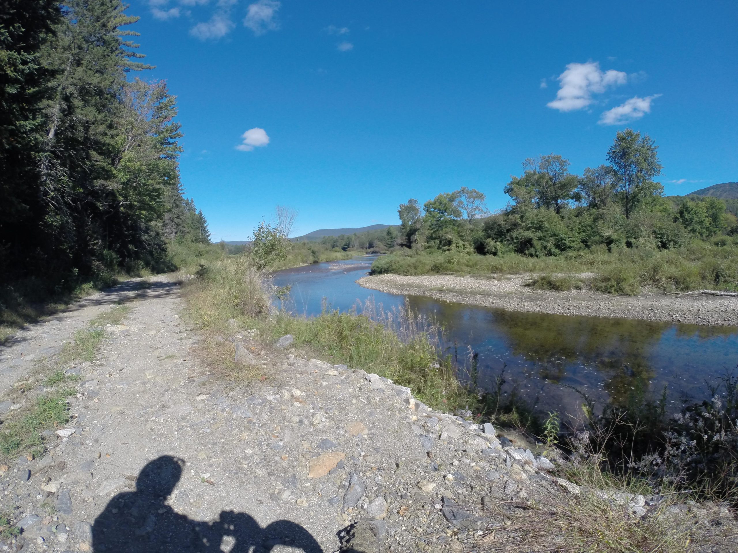 A scenic view of a dirt path lined with greenery alongside a calm river, under a clear blue sky with a few clouds. The surrounding trees and mountains create a tranquil natural setting. A shadow of a person is visible in the foreground, suggesting a moment of exploration or biking. Kingdom Trails mountain bike trail.