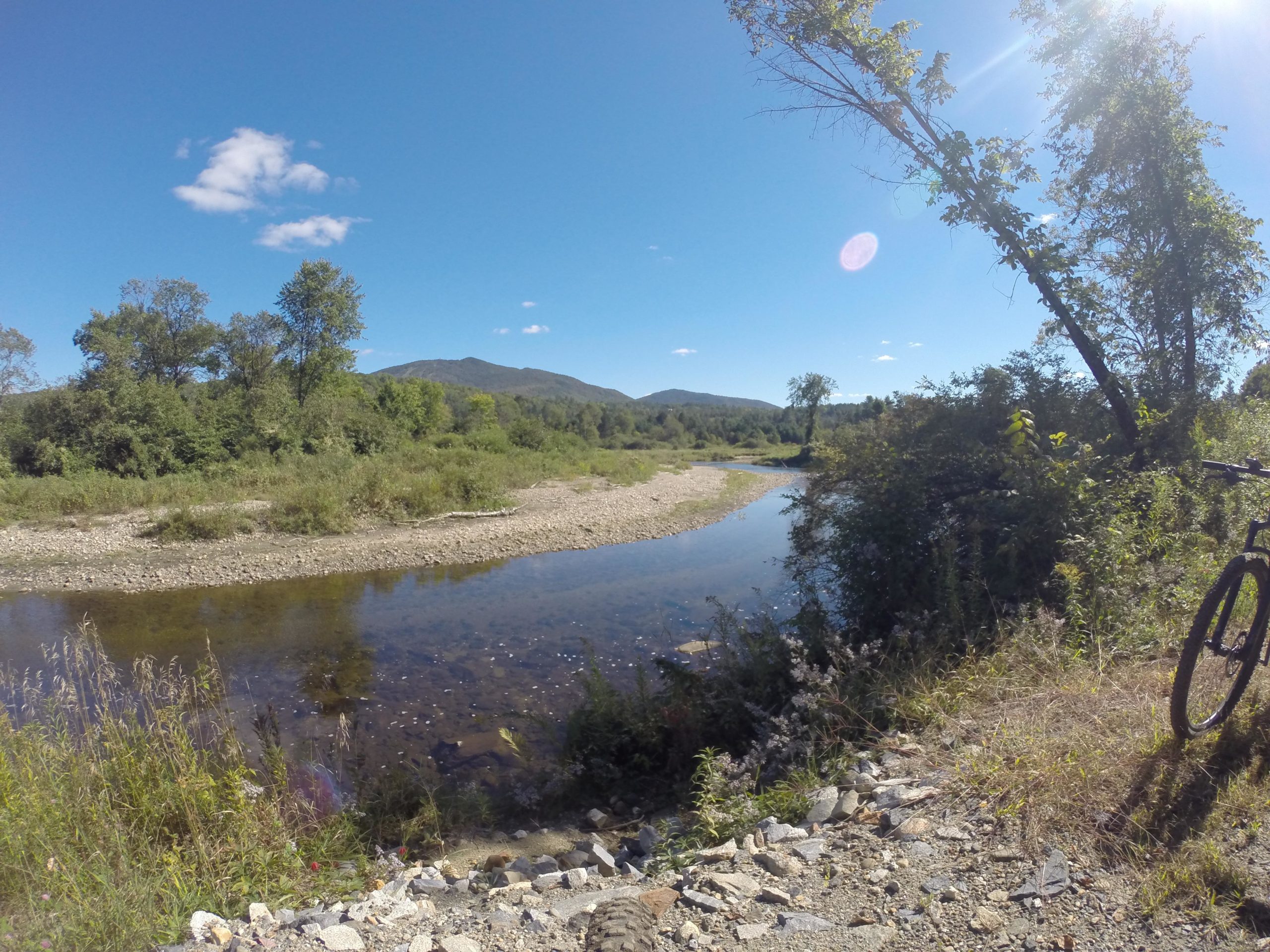 A scenic view of a river winding through lush greenery, with a backdrop of hills under a clear blue sky. The foreground features pebbles and grass along the water's edge, while a bicycle is visible on the right side of the image. Kingdom Trails mountain bike trail.