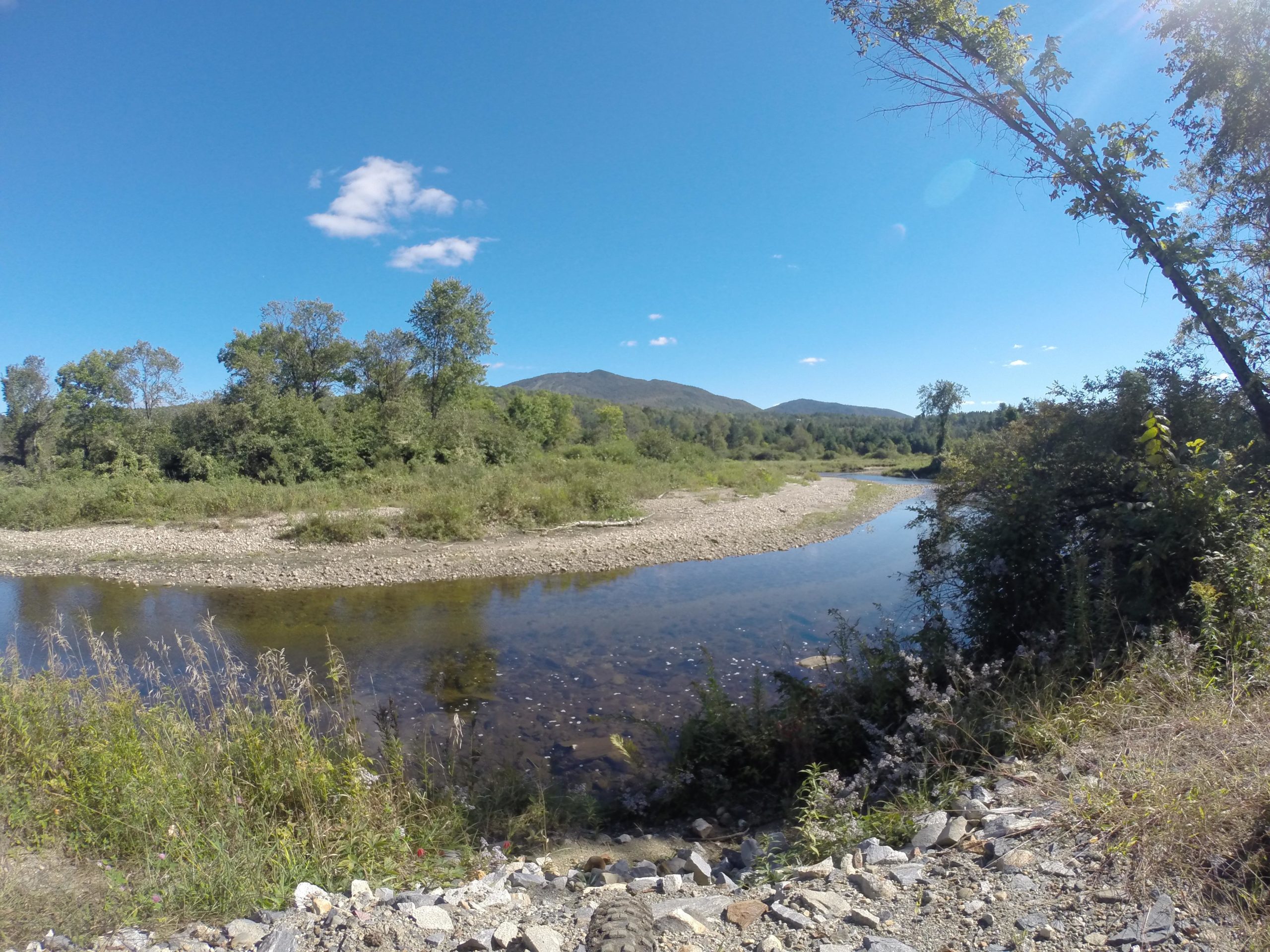 A scenic view of a calm river winding through a lush, green landscape. The riverbanks are lined with trees and bushes, with rocky areas visible along the water's edge. In the background, hills rise under a clear blue sky dotted with a few fluffy clouds. The scene depicts a tranquil natural setting, perfect for outdoor activities. Kingdom Trails mountain bike trail.