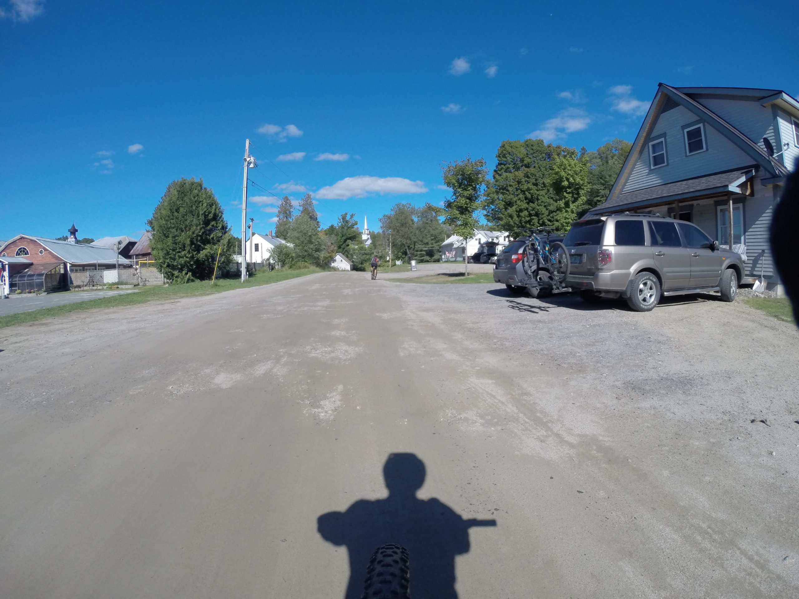 A dirt road stretches ahead with a cyclist riding in the distance. On the right, a driveway features a parked SUV with a bike rack, while a house with a peaked roof is in the foreground. Lush trees and buildings line the road under a clear blue sky with a few scattered clouds. Kingdom Trails mountain bike trail.