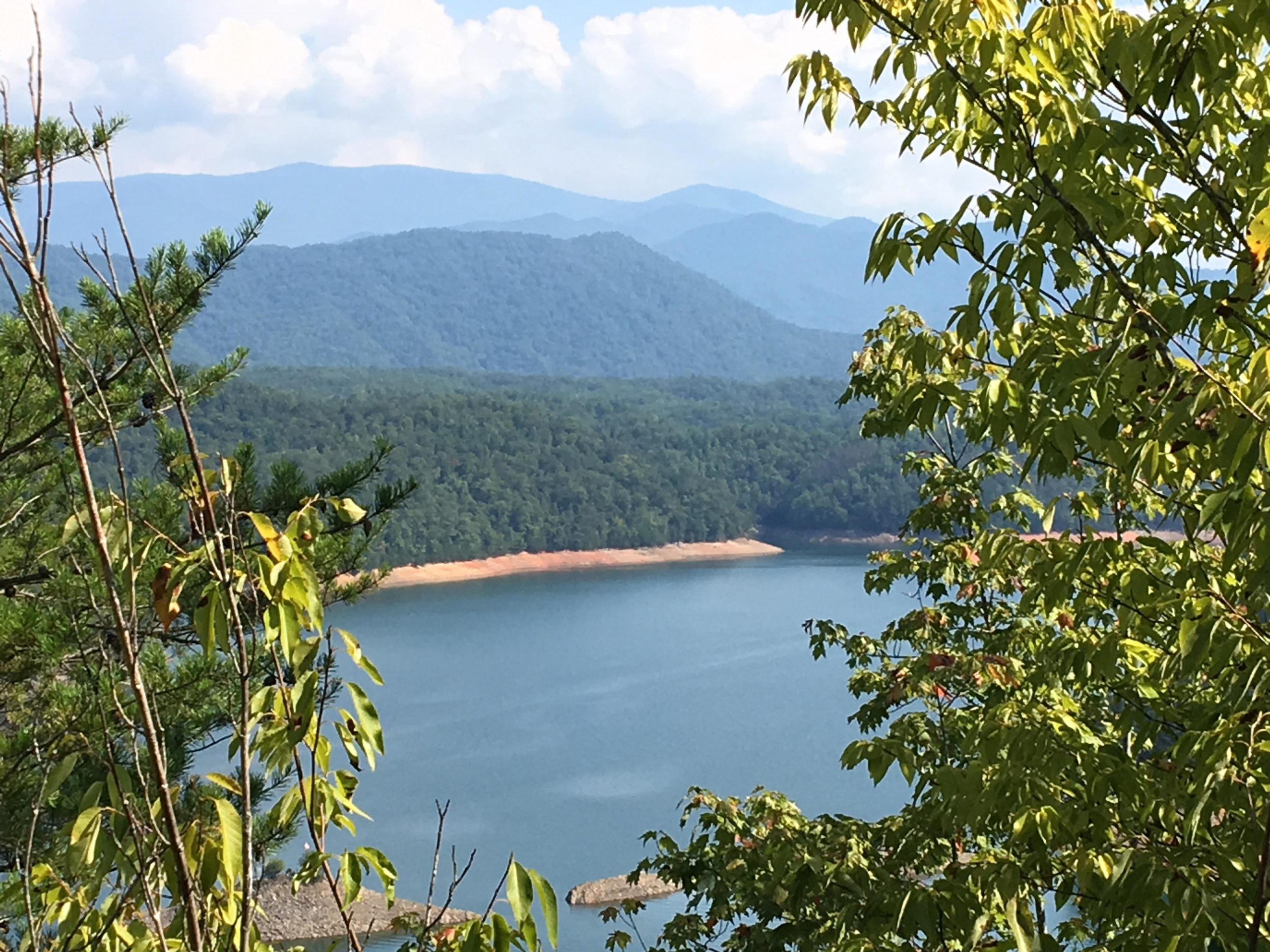 A serene landscape view featuring a tranquil lake surrounded by lush green trees, with rolling mountains in the background under a partly cloudy sky. Tsali Thompson Loop mountain bike trail.