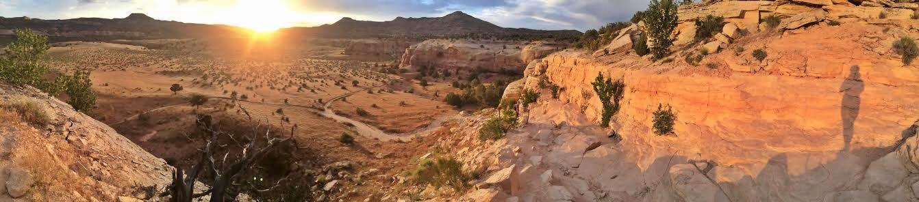 A panoramic view of a desert landscape at sunset, showcasing warm, glowing hues on rocky cliffs and sparse vegetation. Silhouettes of shrubs and trees are visible against the vibrant sky, with rolling hills in the background. A person's shadow is cast on the rocky surface in the foreground. Mary's Loop / Horsethief Bench mountain bike trail.